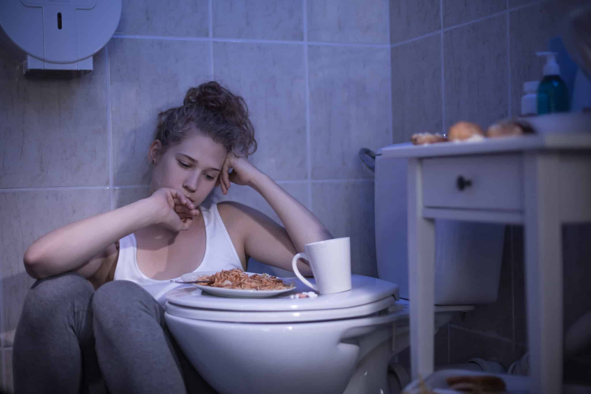 Image of a distressed woman in the bathroom, staring at food in the toilet, symbolizing the struggles and emotional distress associated with bulimia.